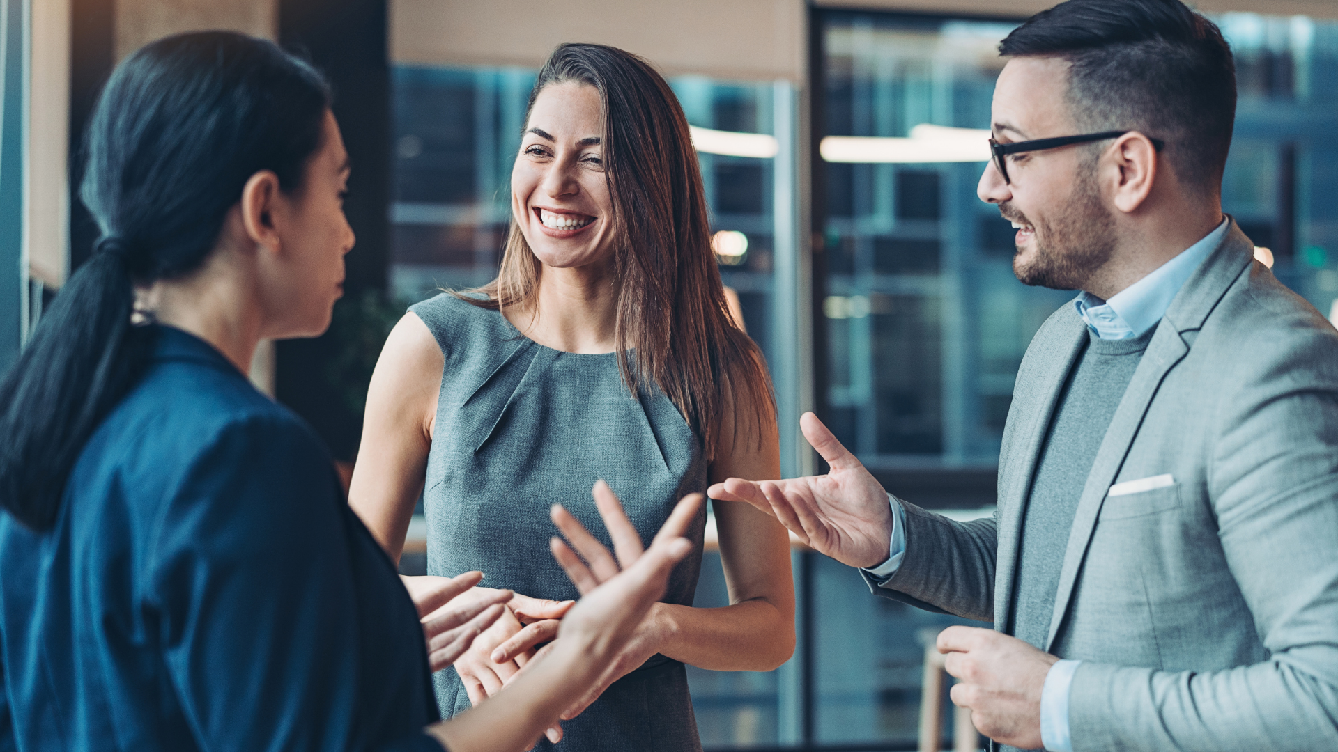 Drei junge Erwachsene in Businesskleidung stehen in einem modernen Büro mit Glasfassade und lachen herzlich bei einem Gespräch. Die freundschaftliche Atmosphäre und lebendige Gestik deuten auf ein positives Arbeitsklima und vertrauensvollen Austausch im beruflichen Kontext hin.