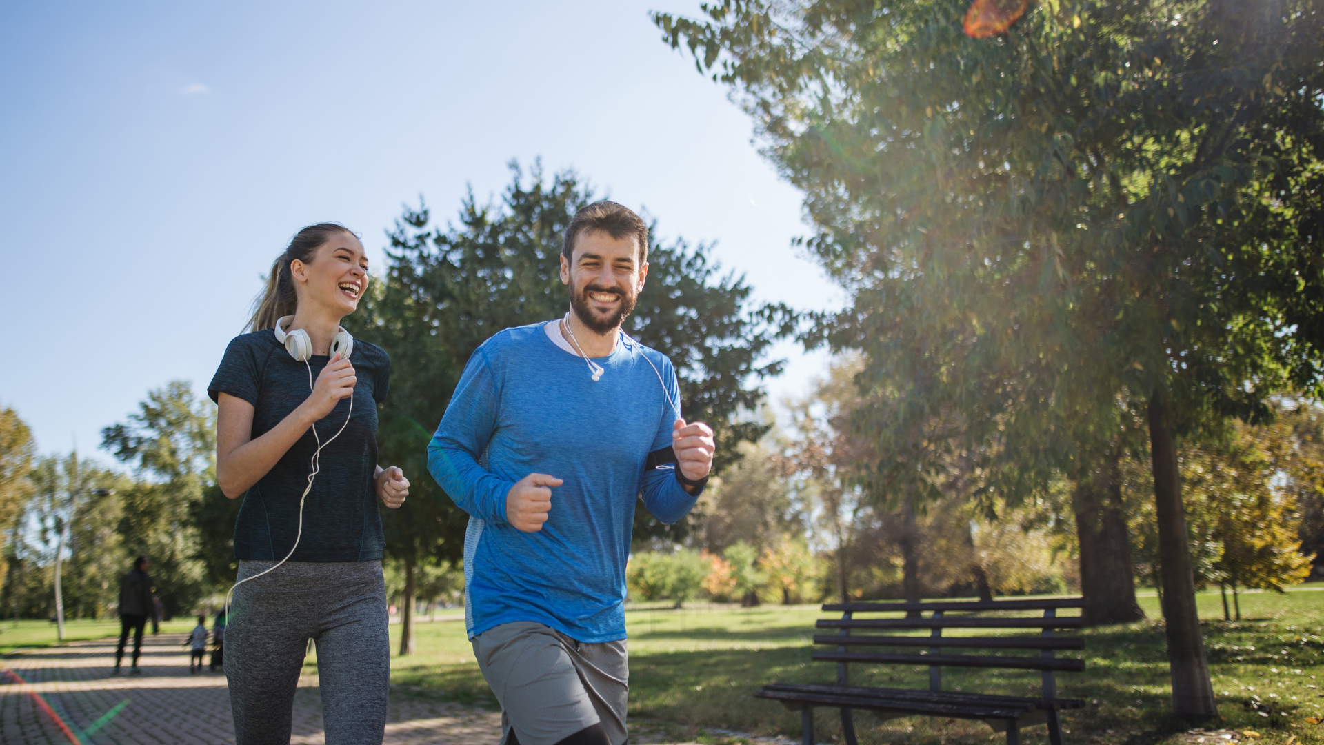Ein Mann und eine Frau joggen lachend nebeneinander auf einem Weg in einem sonnigen Park. Die Szene zeigt Gesundheit, Bewegung und Lebensfreude.
