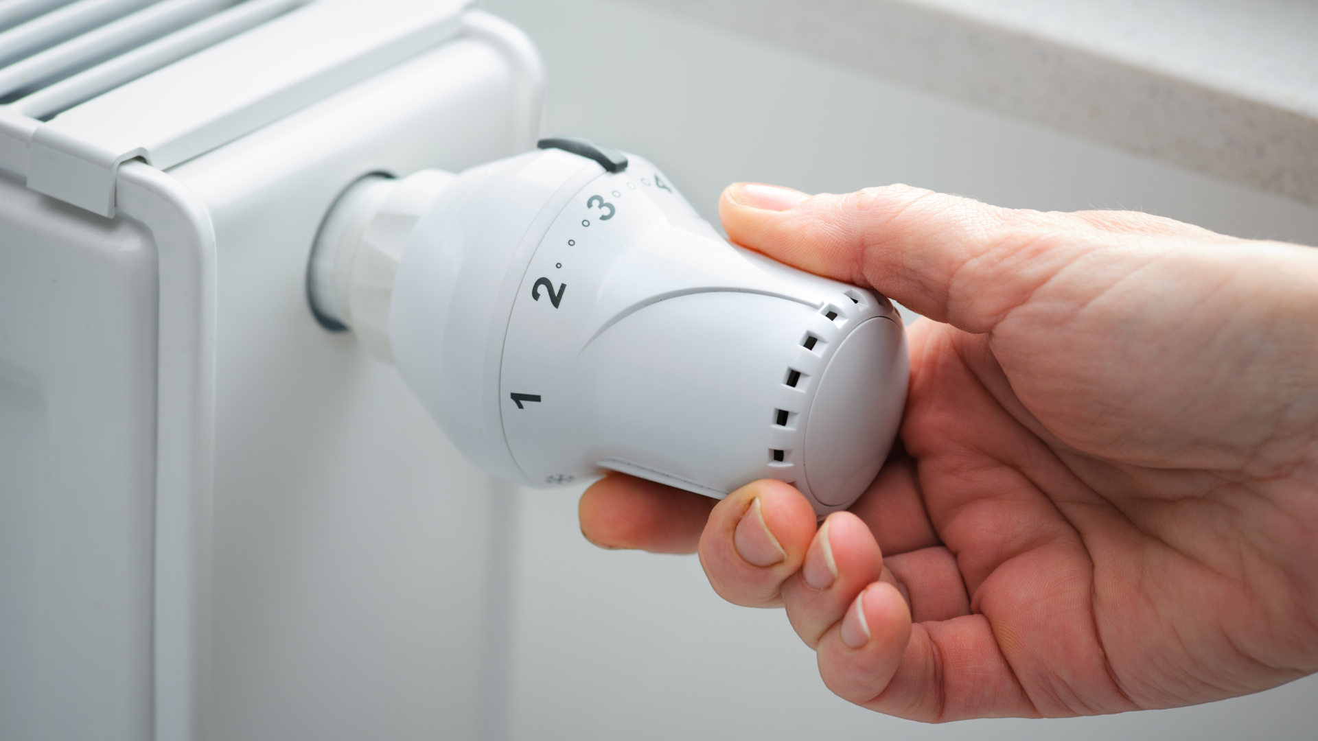 Das Bild zeigt eine Hand, die an einem Thermostat eines Heizkörpers dreht. Der Thermostat ist auf eine niedrigere Einstellung eingestellt, wahrscheinlich um die Raumtemperatur zu regulieren oder Energie zu sparen. Die Szene spielt sich in einem gut beleuchteten Raum ab, was darauf hinweist, dass es sich um eine alltägliche Haushaltshandlung handelt. Das Drehen des Thermostats ist eine einfache, aber wichtige Maßnahme zur Kontrolle des Energieverbrauchs in einem Haushalt.