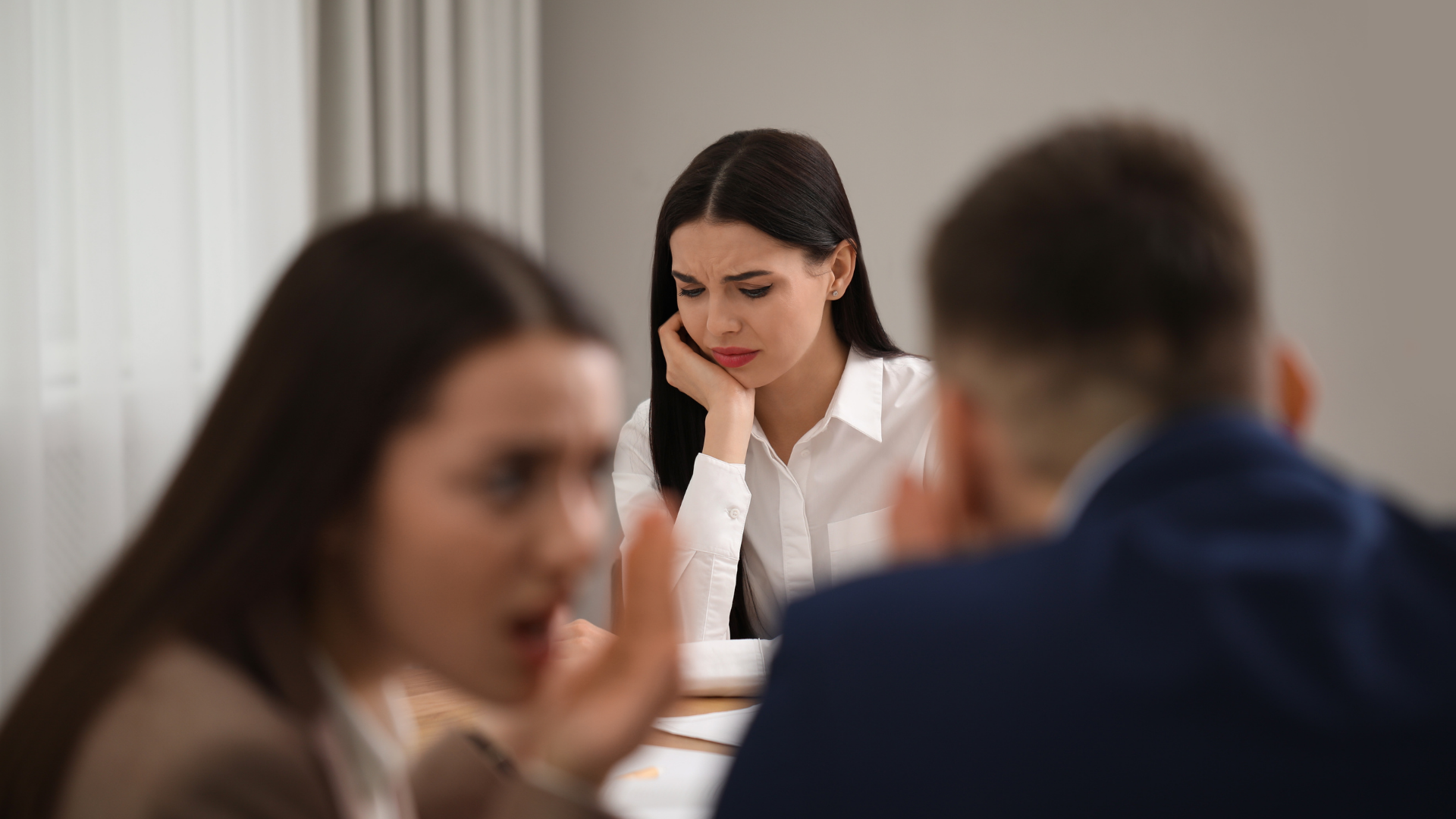 Das Bild zeigt eine Büroszene mit drei Personen, die offenbar in eine ernste Diskussion verwickelt sind. Eine junge Frau im Vordergrund wirkt besorgt und gestresst, während sie ihr Gesicht mit der Hand stützt. Im Hintergrund sind zwei Kollegen zu sehen, wobei einer von ihnen sichtbar gestikuliert und möglicherweise in einem hitzigen Gespräch ist. Die Szene könnte auf ein schwieriges Arbeitsumfeld oder eine Konfliktsituation im Büro hinweisen, bei der die Frau sich möglicherweise ausgegrenzt oder unter Druck gesetzt fühlt. Der Fokus auf ihre besorgte Mimik betont die emotionale Intensität des Moments.