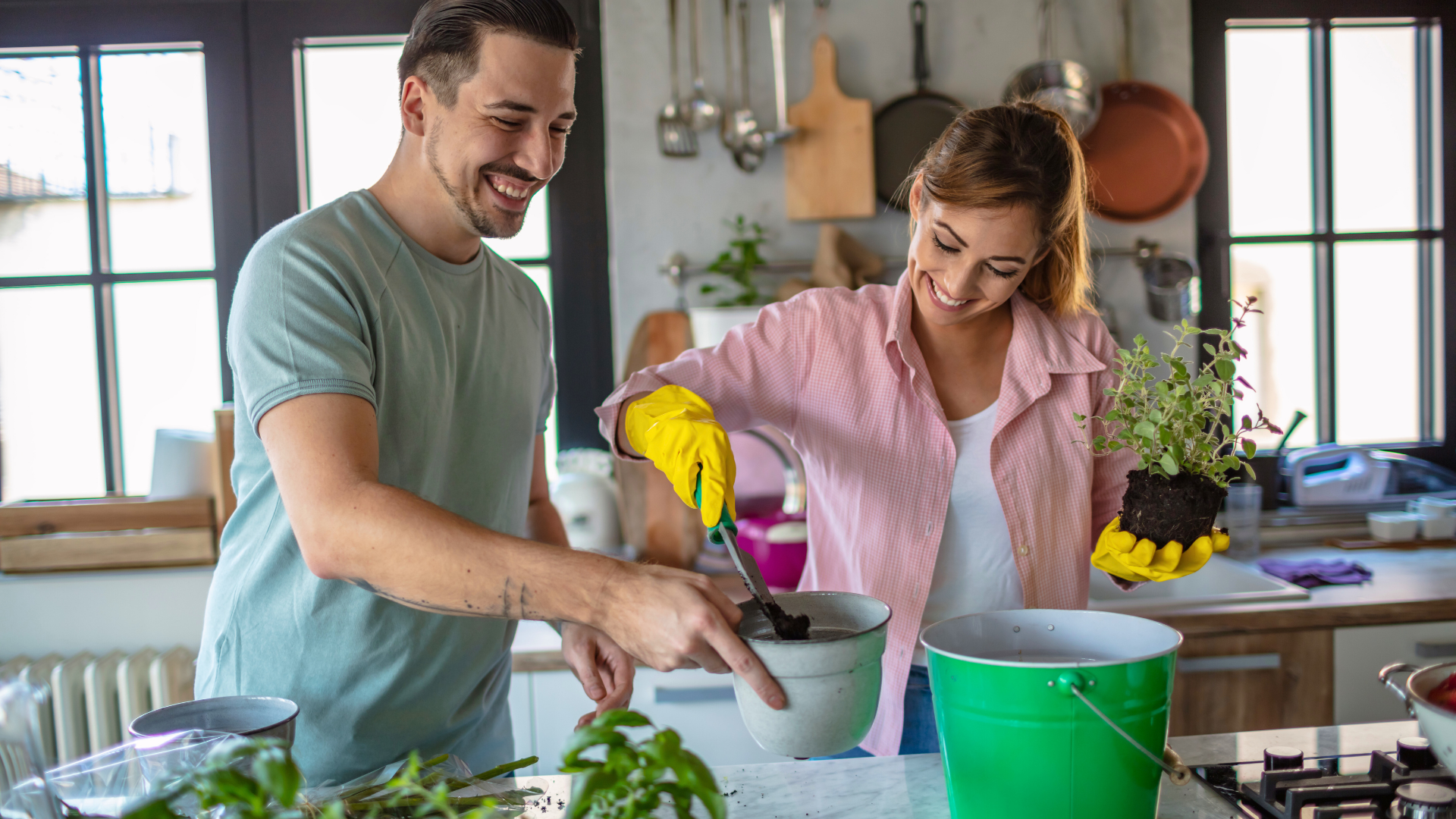 Ein junges Paar steht lachend in einer modernen Küche und topft gemeinsam Kräuter um. Die Szene zeigt Nachhaltigkeit, Gemeinschaft und aktives Gestalten im Alltag.
