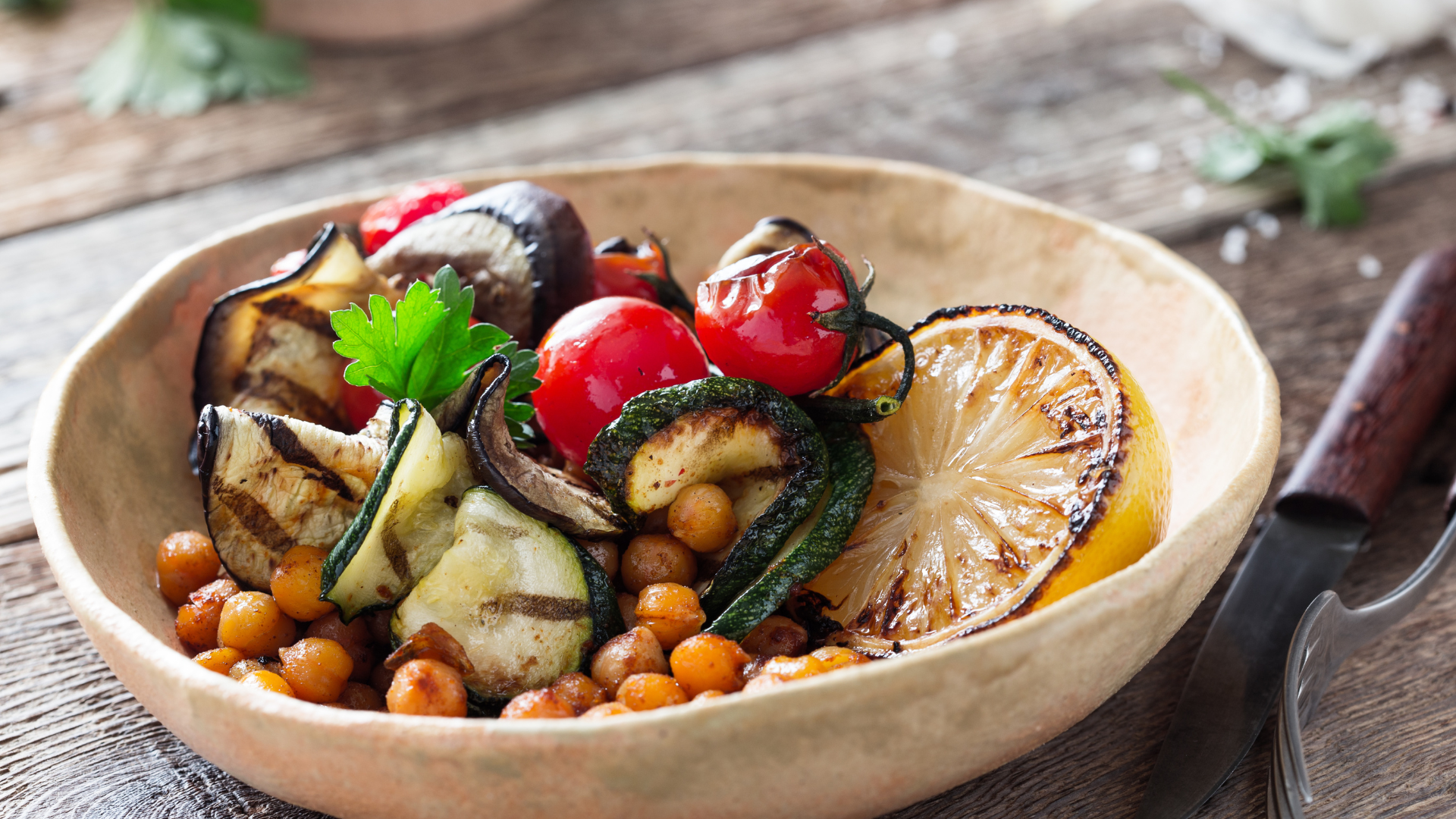Das Bild zeigt eine Schüssel mit gegrilltem Gemüse. Zu sehen sind Zucchini, Auberginen, Kichererbsen und Kirschtomaten, die appetitlich angerichtet sind. Einige der Gemüsestücke haben deutliche Grillspuren. Ein Zitronenschnitz, ebenfalls gegrillt, liegt im Vordergrund, was eine frische Note hinzufügt. Das Ganze wird mit einem frischen Kräuterblatt garniert und auf einem rustikalen Holztisch präsentiert, neben dem ein Messer und eine Gabel liegen.