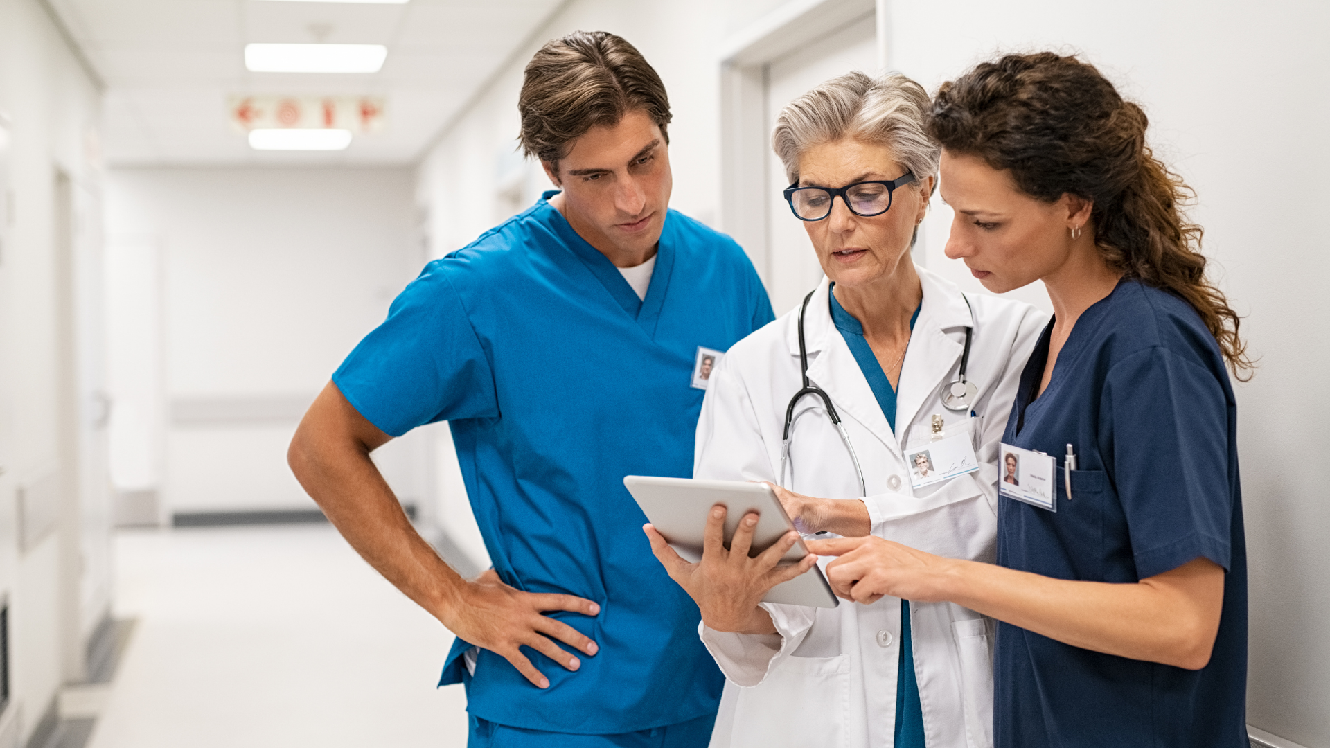 Das Bild zeigt drei medizinische Fachkräfte – zwei Frauen und einen Mann – in einem Krankenhausflur. Sie betrachten gemeinsam Informationen auf einem Tablet, das die ältere Ärztin in den Händen hält. Sie trägt eine weiße Kitteljacke und Brille, während ihre Kollegen in blauen OP-Kleidung gekleidet sind. Die Szene deutet auf eine Beratung oder Diskussion über Patientendaten oder Behandlungspläne hin. Alle Personen scheinen konzentriert und engagiert zu sein, was auf die Wichtigkeit der besprochenen Informationen hindeutet. Dies könnte ein typisches Szenario in einem Krankenhaus sein, wo Teamarbeit und der Austausch von Fachwissen entscheidend sind.
