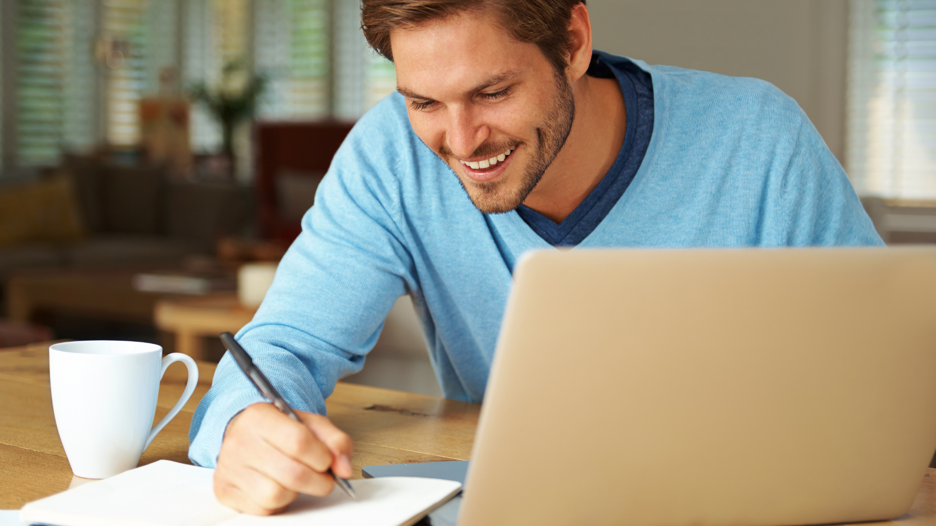 Ein junger Mann sitzt in einem wohnlichen Umfeld mit Laptop und Kaffeetasse am Tisch und schreibt mit einem Stift in ein Notizbuch. Er wirkt motiviert und zufrieden. Die Szene illustriert die Nutzung eines digitalen Ratgebers oder die selbstständige Vorbereitung auf ein Thema, etwa im Bereich Verbraucherbildung.