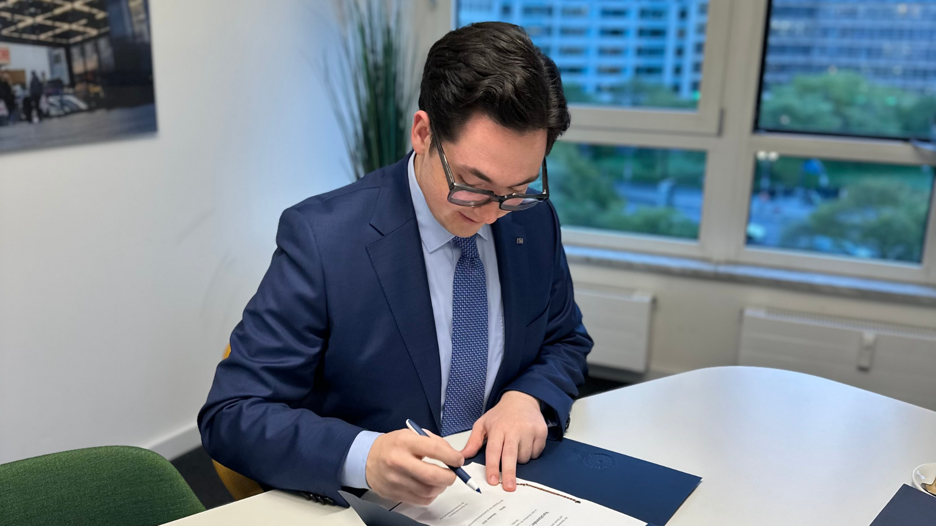 Ein junger Mann im blauen Anzug und gemusterter Krawatte sitzt in einem Büro mit großem Fenster. Er beugt sich konzentriert über eine Mappe und unterzeichnet ein offizielles Dokument. Im Hintergrund ist die Berliner Skyline zu erkennen.