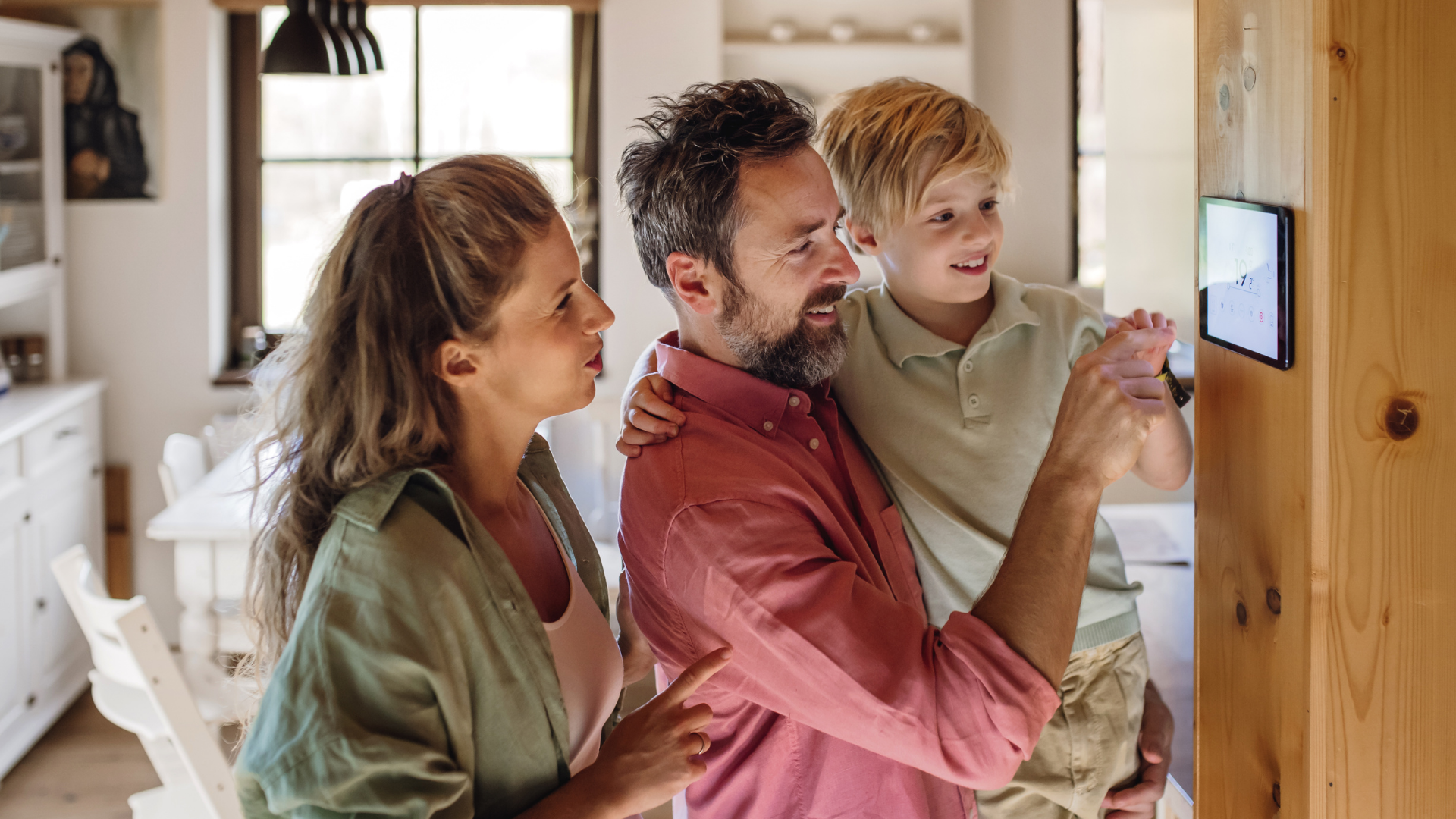 Eine Familie steht in einem hellen, modernen Wohnraum vor einem an der Wand montierten Smart-Home-Bedienfeld. Der Vater bedient lächelnd den Bildschirm, der Sohn sitzt auf seinem Arm und schaut interessiert zu. Die Mutter steht daneben und deutet ebenfalls auf das Display. Alle wirken entspannt und fröhlich.