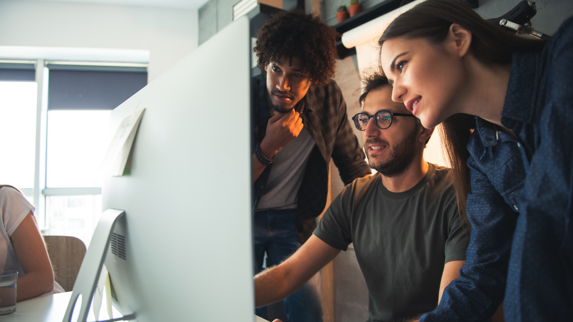 Drei junge Erwachsene arbeiten gemeinsam an einem großen Bildschirm in einem modernen Büro. Eine Frau lehnt sich interessiert nach vorne, ein Mann mit Brille bedient die Maus, während ein weiterer Kollege aufmerksam danebensteht. Die Szene strahlt Teamarbeit, kreative Problemlösung und kollaborative Start-up-Atmosphäre aus.
