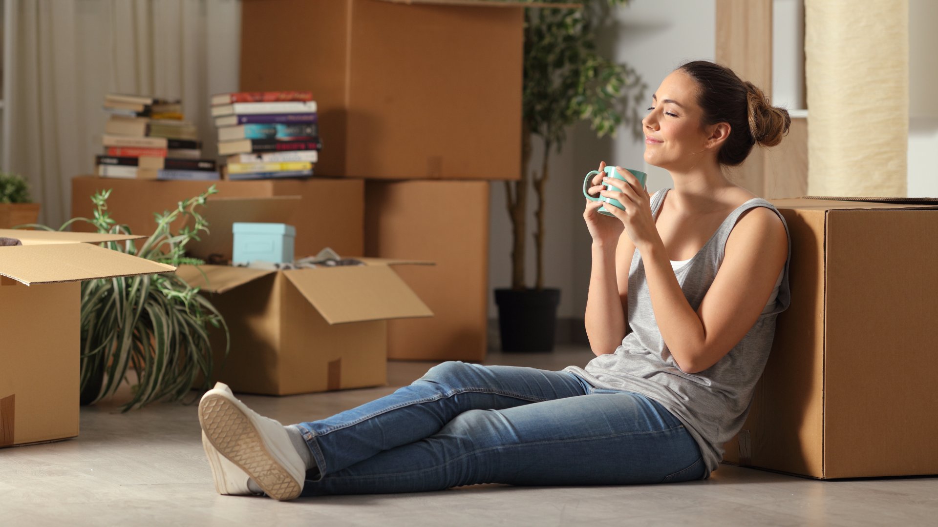Eine junge Frau sitzt entspannt auf dem Boden zwischen Umzugskartons, hält eine Tasse und genießt eine Pause. Die Szene steht für einen Neuanfang oder das Ankommen in den eigenen vier Wänden.