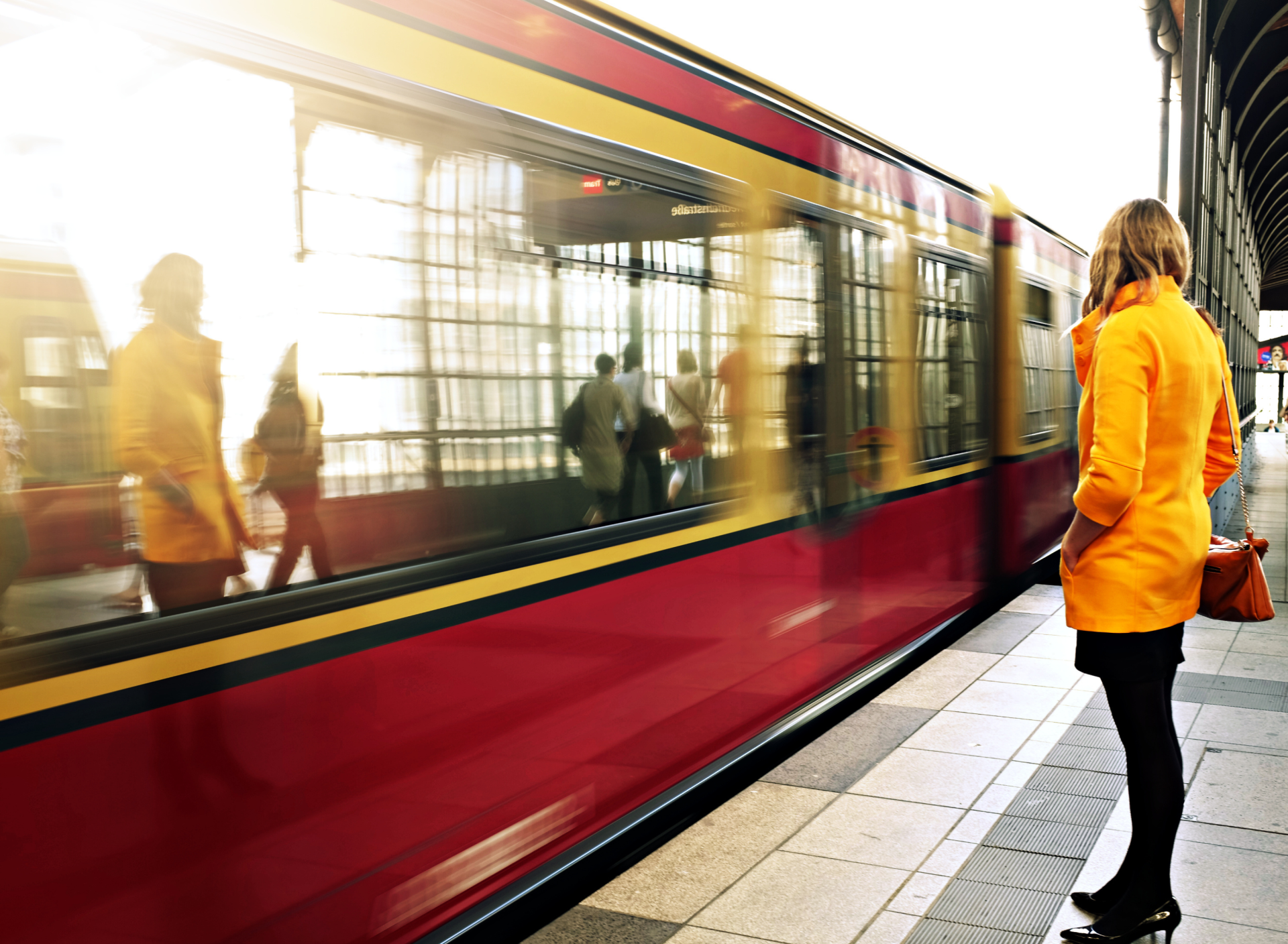 Frau in organgem Kurzmantel steht am Bahnstieg, währen der zug vorbei fährt.