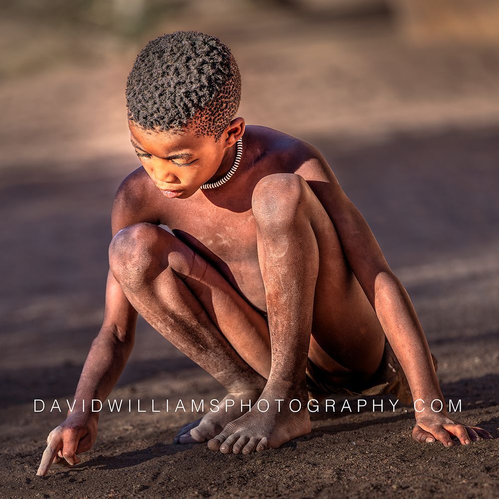 Young Himba boy crouching and drawing with his fingers in the dirt in golden light in Damaraland, Namibia.