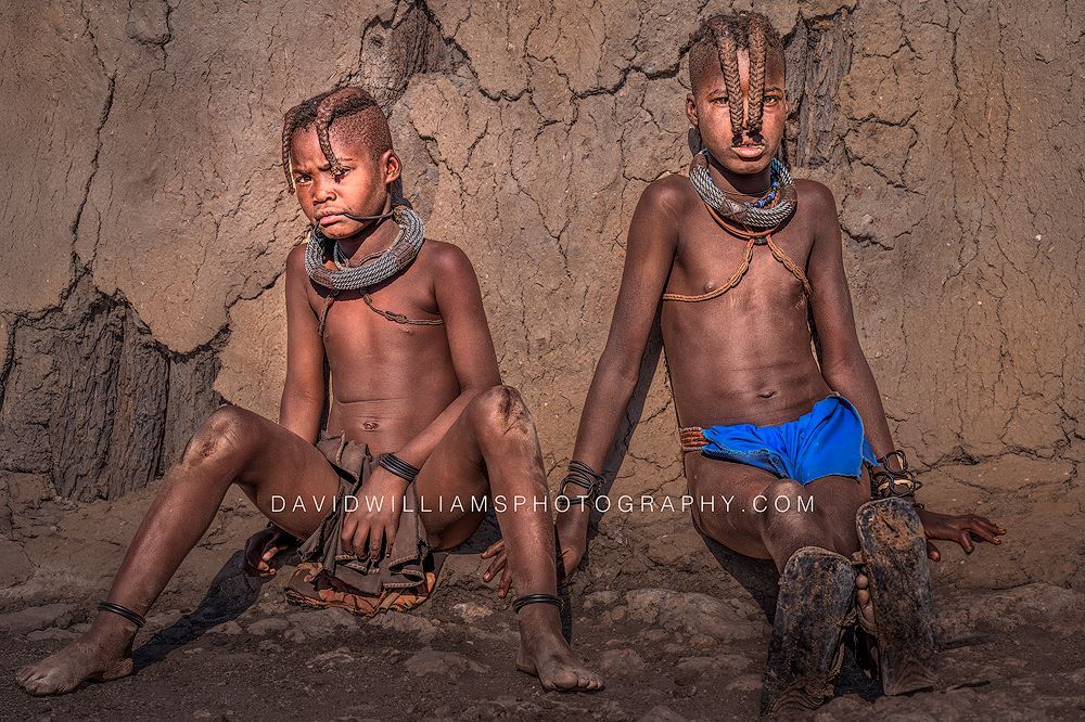 Two teenage Himba boys with braided hair and direct eye contact sitting against their mud home with scowling expressions in Damaraland, Namibia.