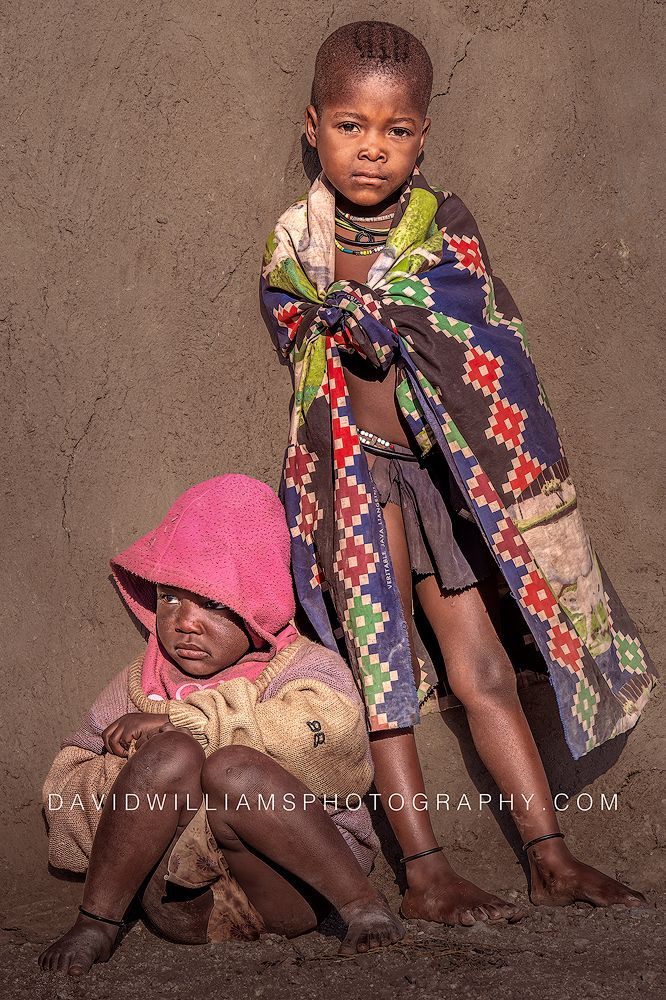 A young Himba boy and girl in weathered clothing lean against their family’s mud home in Damaraland, Namibia.