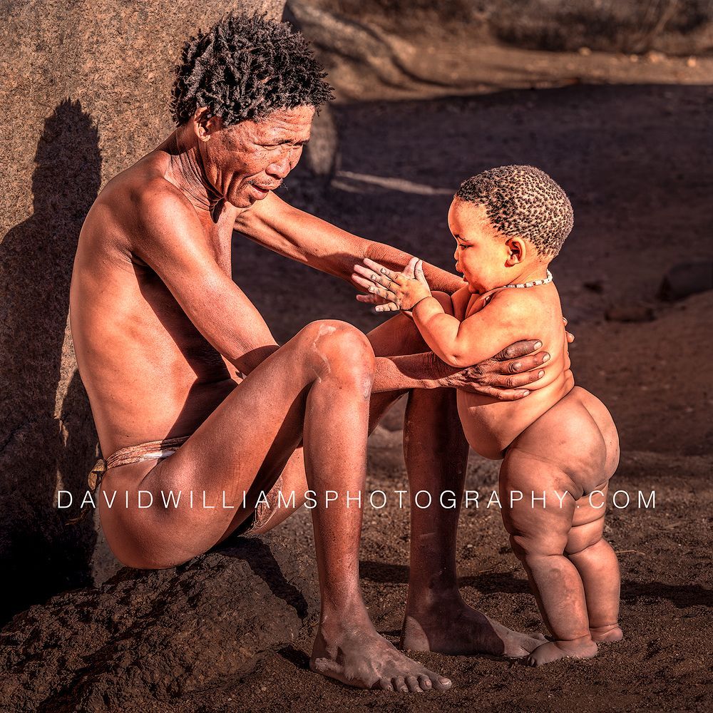 A Himba man plays with and tends his child beside a large rock wall in Damaraland, Namibia.