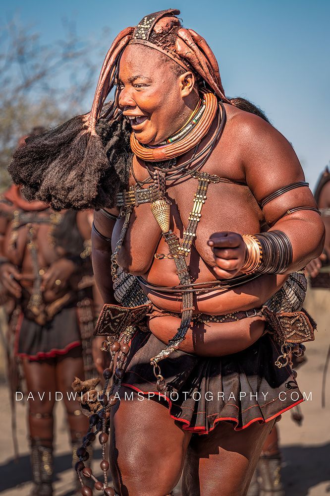 Smiling Himba woman performing the Ondjongo dance of happiness in sunshine with other Himba women in the background in Damaraland, Namibia.