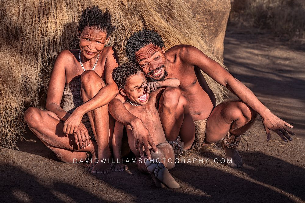 A San Tribe couple sits with their son in front of a grass hut, smiling and laughing in late-day sun near Spitzkoppe, Namibia.
