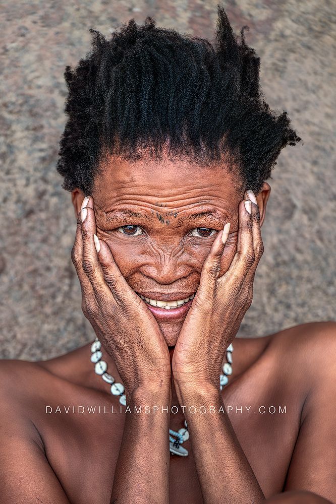 A smiling San Tribe woman poses with her hands on her face in a vertical portrait near Spitzkoppe, Namibia.