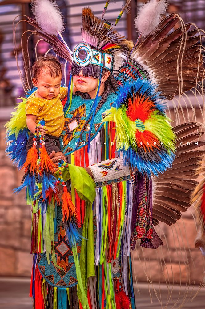 Navajo man in colorful neon regalia holding his young child during a break from a traditional war dance, vibrant and ornate from head to toe
