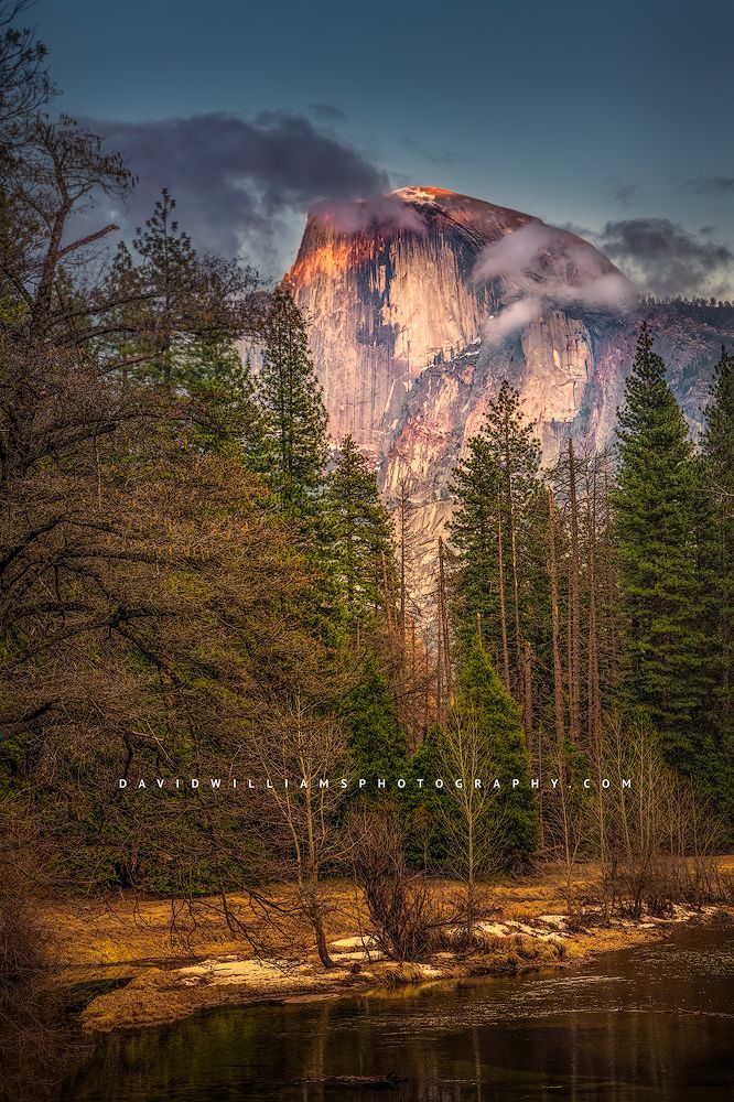 The sun is rising over Half Dome against the green forest trees and Merced River, taken from the Sentinel Bridge viewpoint in Yosemite National Park, California