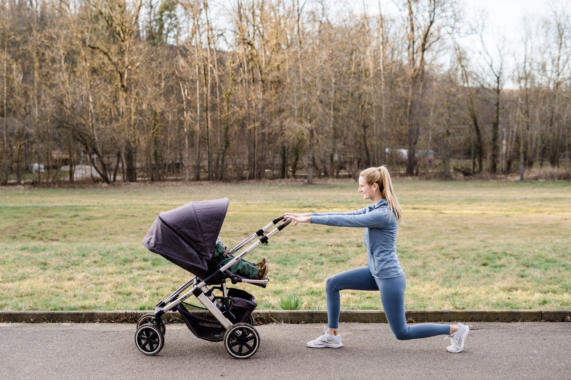 Julez macht Sport mit Baby draußen im Freien. Das Baby sitzt im Kinderwagen und Julez hält sich daran fest um sich abzustützen. Ihr linkes Bein streckt sie seitlich nach oben. Beide haben viel Spaß zusammen beim Sport.