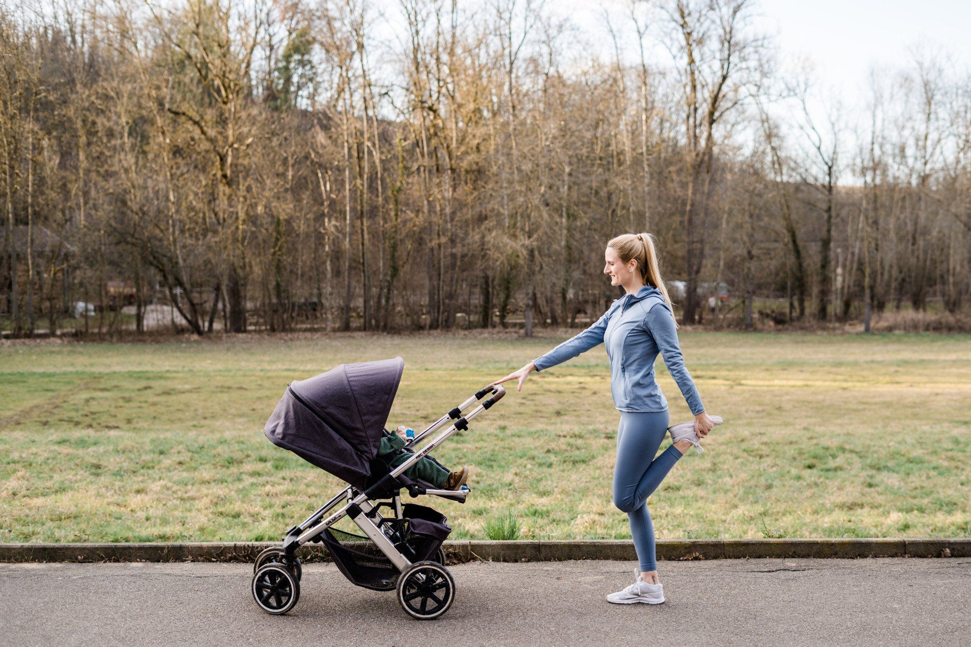 Fit mit Baby Outdoor - Mein Fitnesskurs für alle Mamas mit Baby/ Kind draußen im Freien Hier ist Julez draußen mit dem Kind im Kinderwagen. Sie hält sich am Kinderwagen fest und dehnt ihren Oberschenkel, in dem sie diesen Richtung Po zieht. Sie trägt auch draußen ein blaues Sportoutfit.