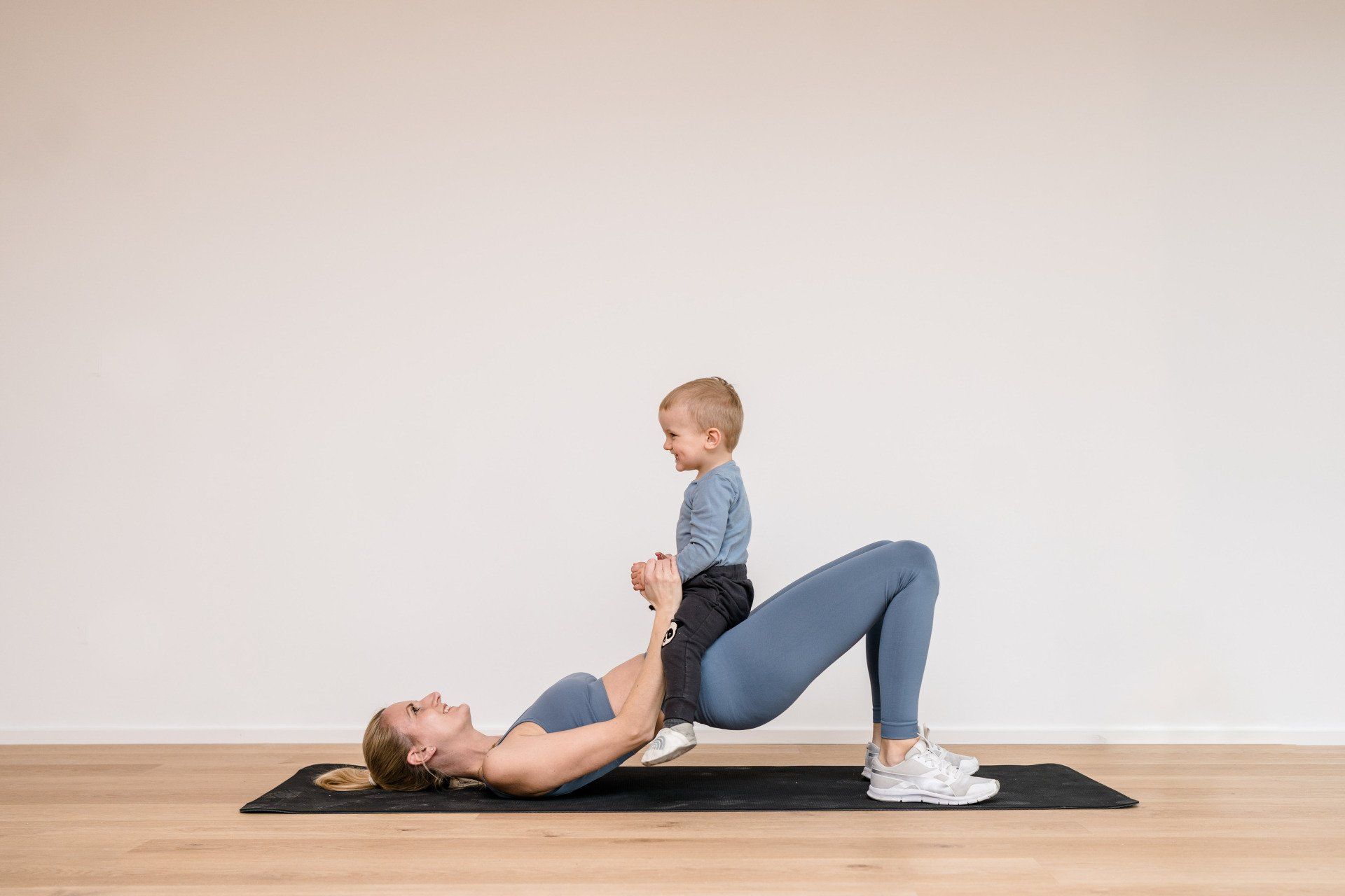 Julez macht Sport mit Baby. Julez liegt mit dem Rücken auf der Sportmatte, die Beine sind angewinkelt und das Baby sitzt auf ihren Hüften, Sie trägt ein blaues Sporttop und eine blaue Hose. Julez und das Baby haben viel Spaß zusammen beim Sport machen.