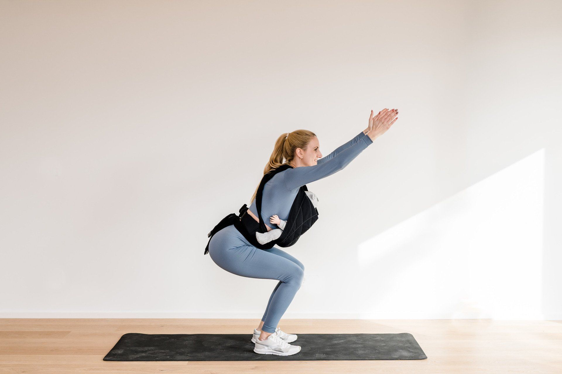 Man sieht hier Julez in ihren blauen Sportklamotten. Sie macht eine Kniebeuge und die Arme sind in die Luft gestreckt. Si9e hat ihr Baby dabei in der Trage.