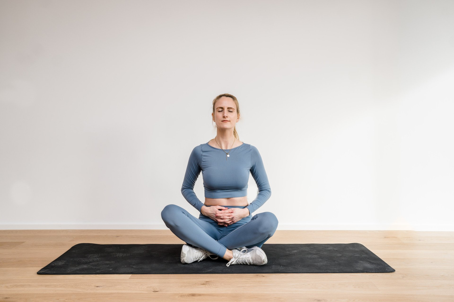 Julez sitzt im Schneidersitz auf der Sportmatte. Ihre Hänge liegen auf Ihrem Bauch und die Augen sind geschlossen. Sie konzentriert sich auf die Atmung.
