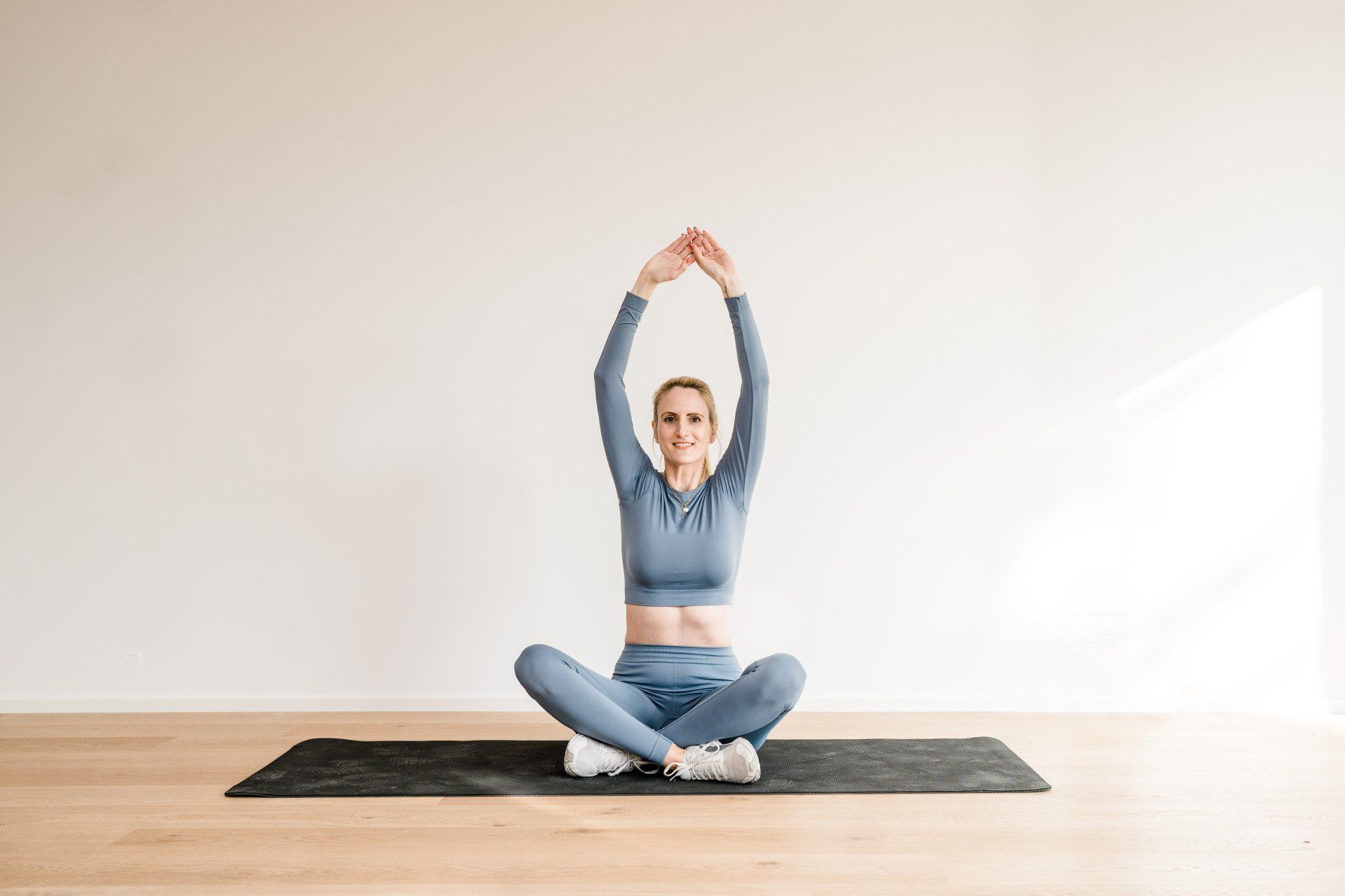 Fit mit Babybauch - Mein Fitnesskurs speziell zugeschnitten für alle schwangeren Frauen Auf dem Bild sitzt Julez im Schneidersitz auf der Sportmatte. Die Arme hält sie nach oben gestreckt, die Finger berühren sich. Der Rücken wird dabei gedehnt. Sie trägt ein blaues Sportoutfit.