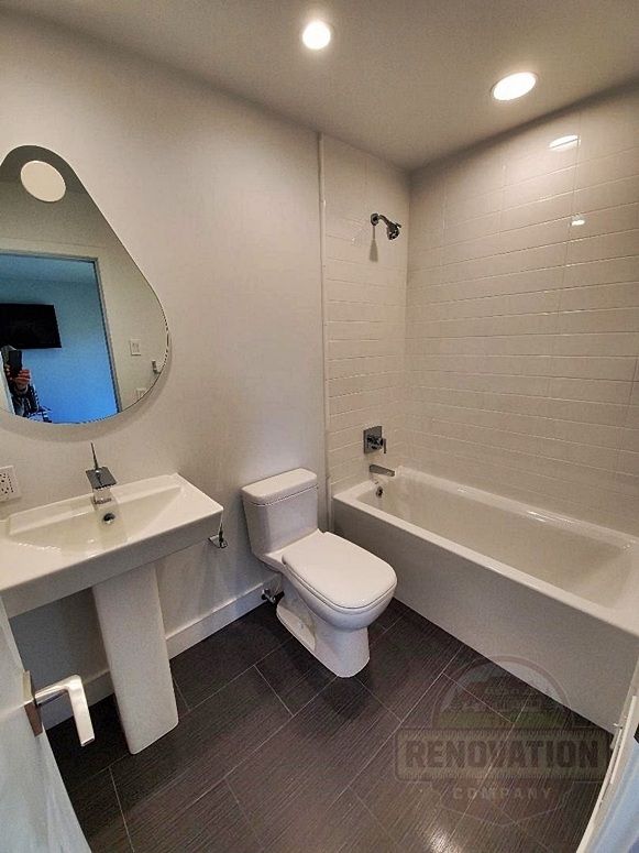 Remodeled full bathroom by The Renovation Company with a pedestal sink, teardrop mirror, white tiled bathtub walls, and a brown tiled floor