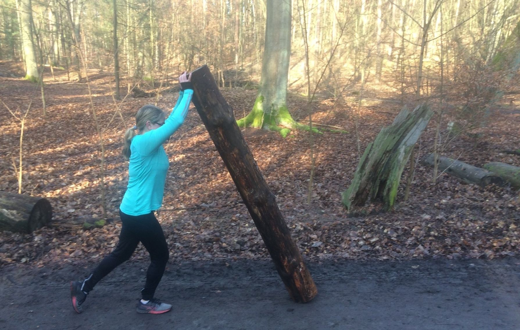 Eine Frau beim Training im Berliner Wald schiebt und hebt einen Baumstamm vor sich her.