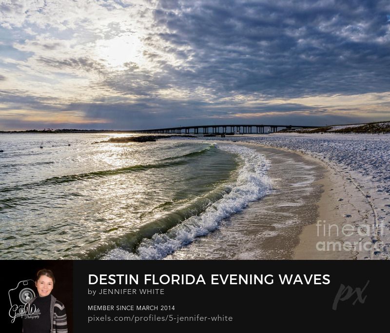 Waves roll in on shore before sunset on a cloudy evening in Destin, Florida, united States. In the distance you can see Destin Bridge and William T. Marler Bridge. You can see a hint of sunrays coming through the clouds as the sun glares on the water which is part of the Emerald Coast and Gulf of Mexico.