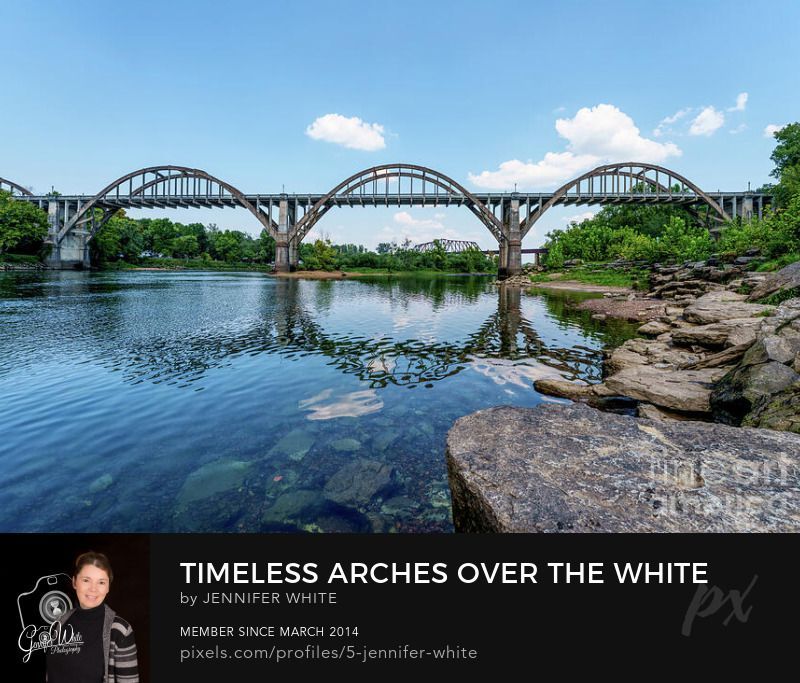 A photograph of the historic Cotter Bridge set along the peaceful banks of the White River in Cotter, Arkansas. It spans the glistening water of the White River with its distinctive concrete arches—a tribute to the craftsmanship of the late 1920s. Completed in 1929 and carefully restored in 2002, this six-span bridge—officially known as the R.M. Ruthven Bridge—shines at night with colorful lights. In the background, a vintage railroad bridge adds a nostalgic layer to the view, stirring echoes of the past. From a viewpoint framed by a weathered rock wall, the scene merges natural textures and historic charm in one timeless composition.