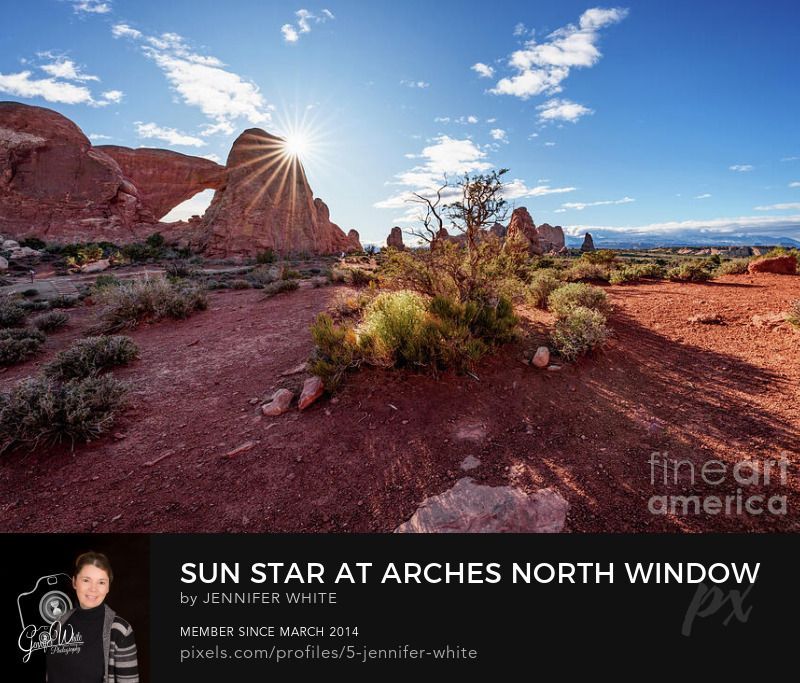 This peaceful early morning photograph at Arches National Park showcases a brilliant sunburst beside the North Window rock formation in the Windows Section near Moab, Utah, United States. The deep orange sandstone of the arch glows with rich, reddish tones as the morning light passes through its natural opening. A few soft clouds drift across the vibrant blue desert sky, while the distant La Sal Mountains add depth to this striking and iconic Utah landscape.
