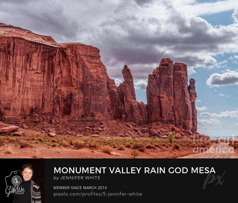 This photograph captures a moody and dramatic view of Rain God Mesa in Monument Valley, Arizona, USA, as storm clouds begin to roll into the area. The scenic drive dirt road winds alongside the towering sandstone formation, drawing the viewer’s eye through the rugged desert landscape. The rich red hues of the mesa contrast beautifully with the darkening gray sky, creating a sense of depth and anticipation as the weather shifts. Subtle light breaks through the clouds, softly illuminating the textured rock face and surrounding desert terrain. This powerful scene showcases the raw beauty and ever-changing atmosphere of Monument Valley, where nature’s artistry and the spirit of the Southwest meet in perfect harmony.