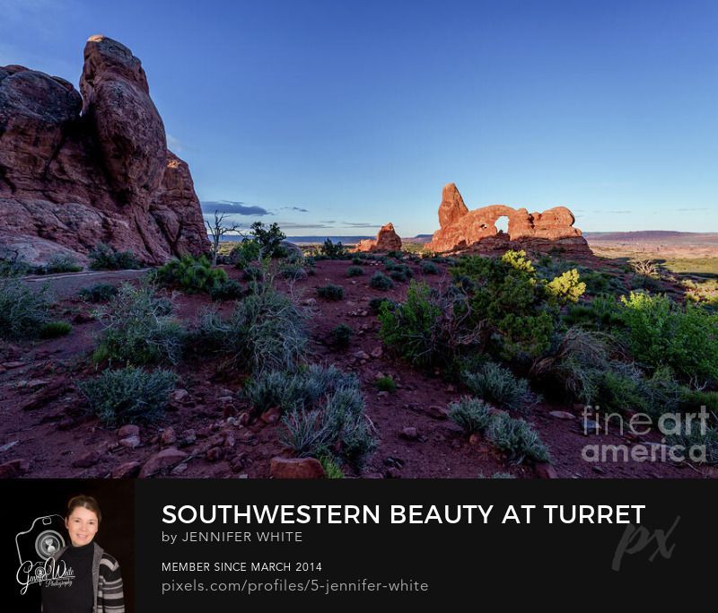 This photograph captures the morning light casting dramatic shadows across Turret Arch in the Windows Section of Arches National Park, Moab, Utah, USA. The rugged sandstone formations rise boldly against a partly cloudy blue sky, where soft light and shadow create depth and contrast. The warm red rock glows orange with the colors of the desert morning, showcasing a peaceful Southwestern scene that reflects the timeless beauty of this iconic landscape.