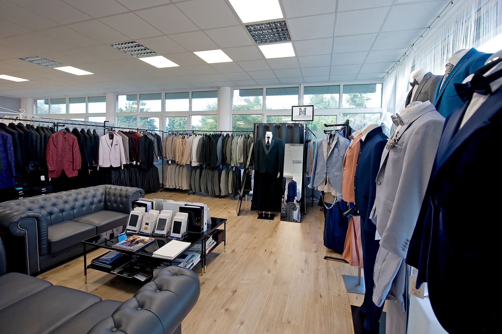 Herrenmodengeschäft mit eleganten Anzügen in verschiedenen Farben und Stilen. Im Vordergrund stehen Schaufensterpuppen mit festlichen Anzügen in Blau, Grau und Beige, während im Hintergrund zahlreiche Anzüge an Kleiderstangen hängen. In der Mitte befindet sich eine gemütliche Beratungsecke mit Ledersofas und Couchtisch.