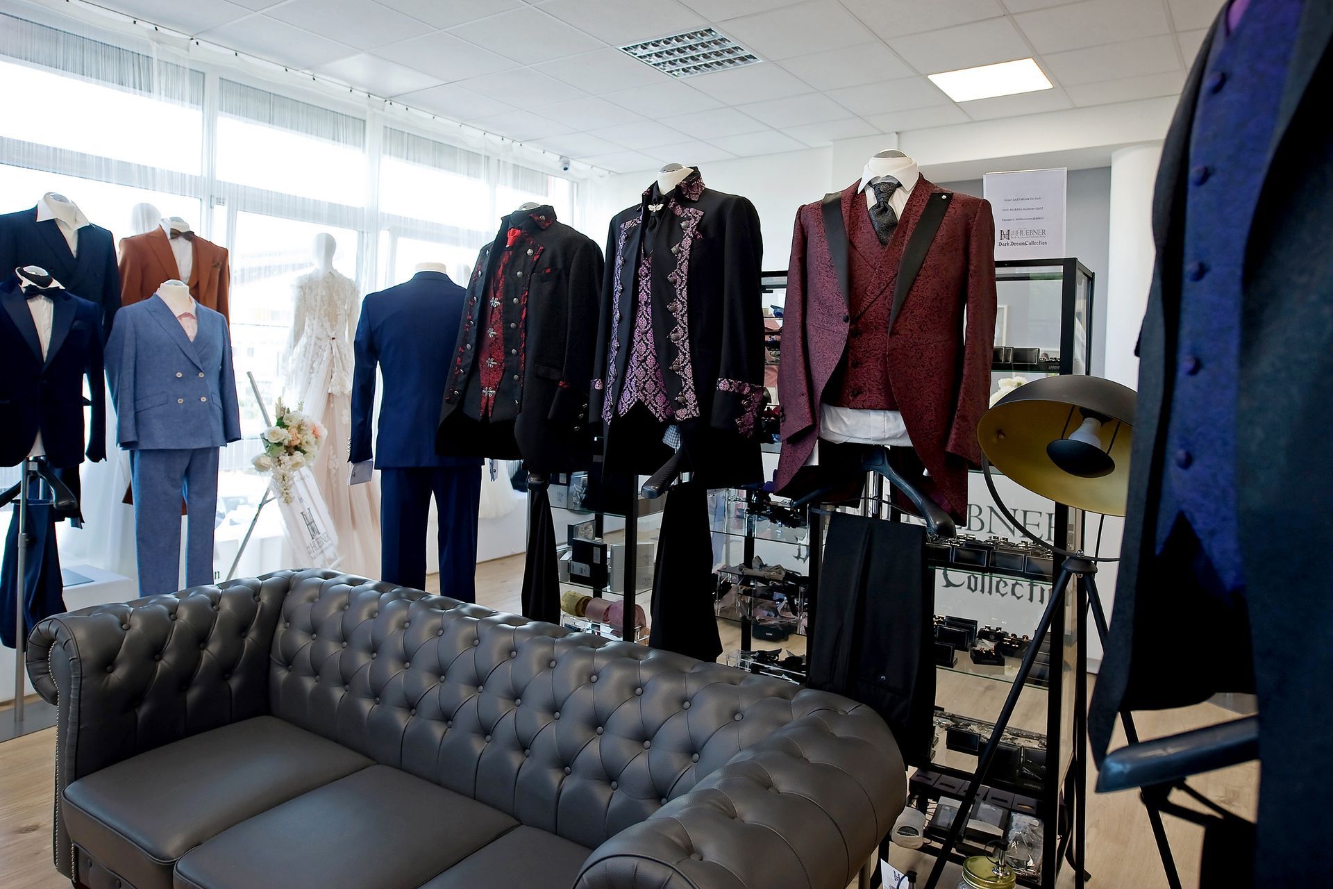 Herrenmodengeschäft mit eleganten Anzügen in verschiedenen Farben und Stilen. Im Vordergrund stehen Schaufensterpuppen mit festlichen Anzügen in Blau, Grau und Beige, während im Hintergrund zahlreiche Anzüge an Kleiderstangen hängen. In der Mitte befindet sich eine gemütliche Beratungsecke mit Ledersofas und Couchtisch