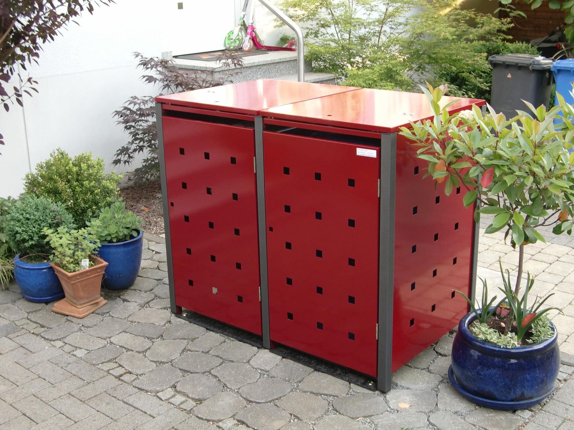 rote Metall Mülltonnenbox für 2 Tonnen steht zwischen Pflanzkübeln