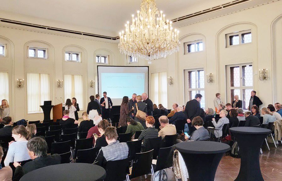 Ein großer Saal mit vielen Stühlen und einer Beamer-Wand im Hintergrund. Viele Menschen sitzen auf den Stühlen oder stehen in den Gängen und unterhalten sich.