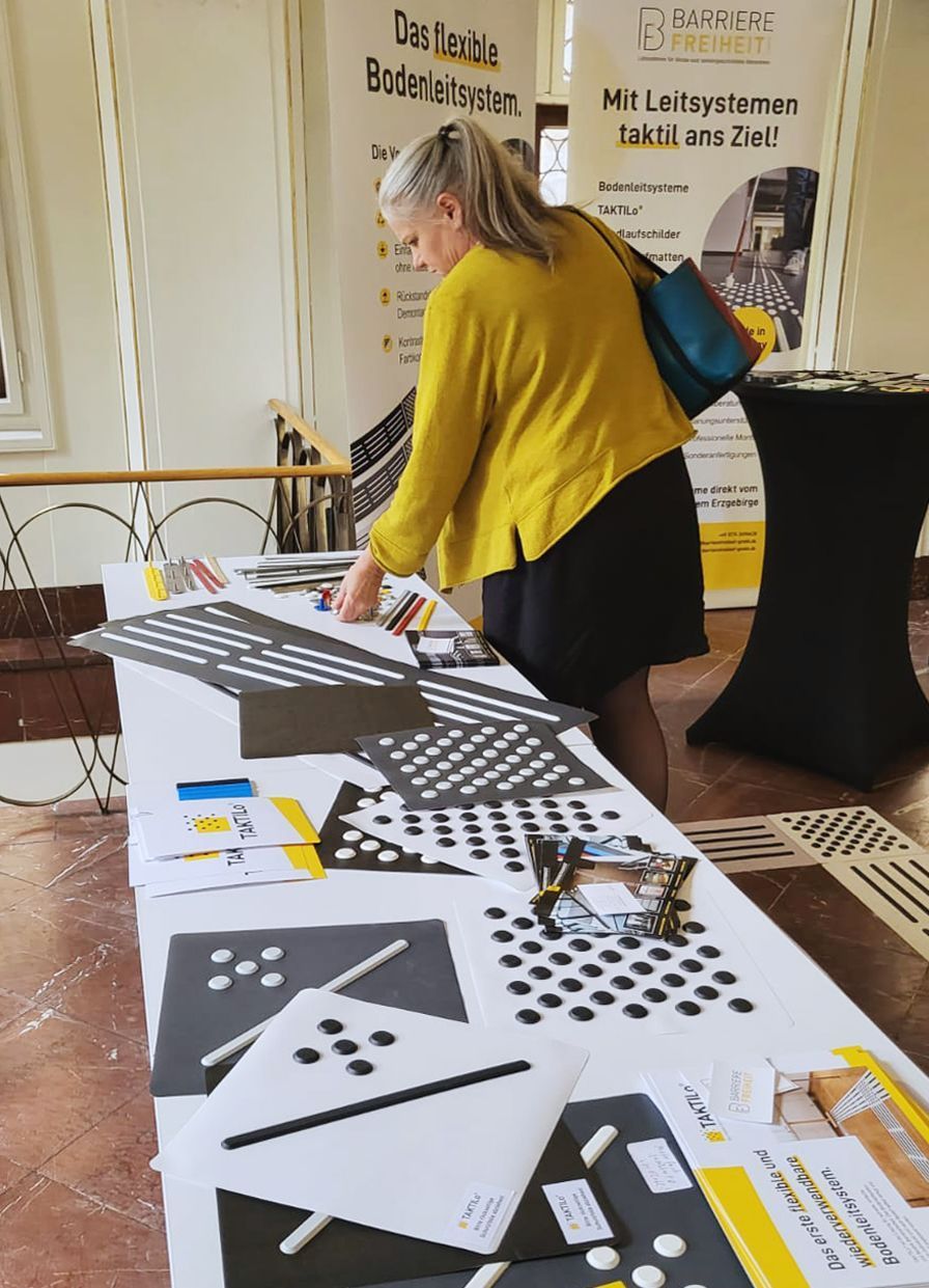 Eine Frau am Ausstelltisch der Barrierefreiheit GmbH. Sie betrachtet die Materialien, die auf dem Tisch ausgelegt sind. Es sind Komponenten eines Bodenleitsystems.