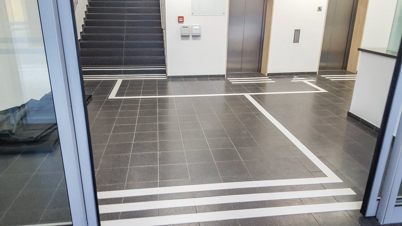Automatische Schiebetüren sind geöffnet und lassen Besucher in den Eingangsbereich eintreten. Ein Bodenleitsystem führt vom Eingang zu zwei Aufzügen und zu einer Treppe