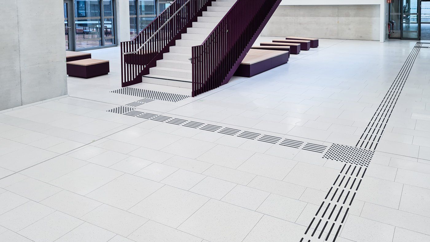 Ein großer Raum mit Treppe und Sitzgelegenheiten. Das Gebäude kann über eine Glastür betreten werden. Von dort aus kann die Treppe über ein Bodenleitsystem erreicht werden.
Entdecken Sie unsere Auswahl an DIN 32984-konformen Bodenleitsystemen, die speziell für die barrierefreie Beschilderung und Orientierungshilfen im öffentlichen Raum entwickelt wurden. Unsere Produkte bieten eine umfassende Palette an taktilem Feedback durch Aufmerksamkeitsfelder, Abzweigefelder und Leitlinien, optimiert für Blinde und Sehbehinderte.
Wir sind stolz darauf, eine breite Auswahl an Bodenindikatoren anzubieten, die nicht nur den aktuellen Standards des barrierefreien Bauens entsprechen, sondern auch die strengen Anforderungen der DIN-Normen erfüllen. Unsere Experten stehen bereit, um Sie bei der Auswahl der passenden Indikatoren zu unterstützen und ein Blindenleitsystem zu planen, das optimal auf Ihre Bedürfnisse abgestimmt ist.
Unsere Bodenleitsysteme, einschließlich ertastbarer Bodeninformationen wie Leitlinien sowie Aufmerksamkeits- und Abzweigefelder, sind so konzipiert, dass sie mit einem Langstock oder durch Fußkontakt wahrgenommen werden können. Dies fördert die Zugänglichkeit und Inklusion in jeder Umgebung und unterstützt die Orientierung von Menschen mit Sehbehinderungen gemäß der DIN 32984 „Bodenindikatoren im öffentlichen Raum“.
Vertrauen Sie auf unsere Expertise in taktile Orientierungshilfen, um eine sichere und inklusive Umgebung für alle zu schaffen.