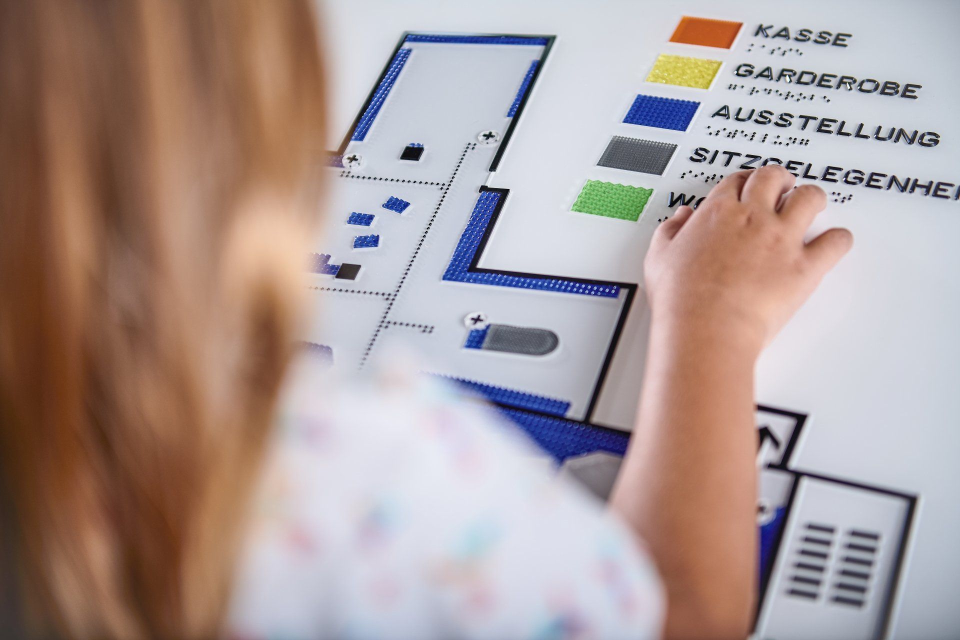 Abbildung einer Person, die vor einem taktilen Grundrissplan für Barrierefreiheit steht und das Wort 'Sitzgelegenheiten' in Brailleschrift ertastet. Dieser Plan bietet einen umfassenden Überblick über die barrierefreie Gestaltung im Gebäude, einschließlich wichtiger Elemente wie Kasse, Garderobe, Ausstellungsbereiche und das integrierte taktile Blindenleitsystem. Jedes Element des Plans ist durch eine einzigartige Haptik gekennzeichnet, die visuell und taktil unterscheidbar ist, was die Orientierung für blinde und sehbehinderte Menschen wesentlich erleichtert und die Zugänglichkeit in öffentlichen Räumen verbessert.