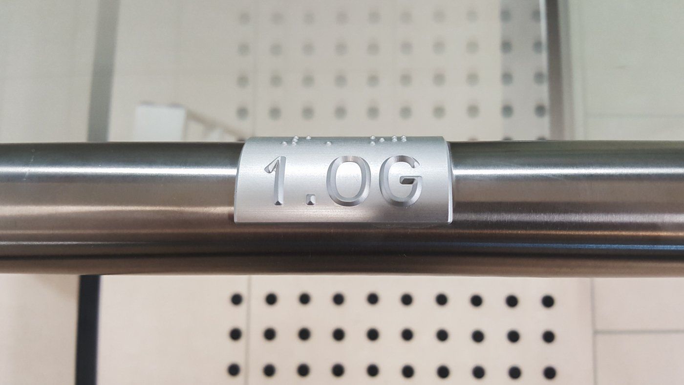 Ein silbernes Handlaufschild mit der Aufschrift 1. OG an einem Geländer. Die Aufschrift ist zusätzlich in Brailleschrift übersetzt und befindet sich Oberhalt der lateinischen Buchstaben.