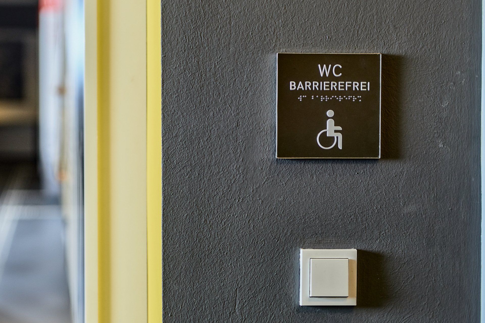 Dieses Bild zeigt ein taktil wahrnehmbares Wandschild für ein barrierefreies WC, gekennzeichnet durch den Text 'WC Barrierefrei' in klar lesbarer Schrift und Braille-Übersetzung. Unterhalb des Textes ist das international anerkannte Piktogramm für Rollstuhlfahrer abgebildet, was die Zugänglichkeit für Menschen mit Mobilitätseinschränkungen hervorhebt. Das Schild unterstützt die Inklusion und Orientierung in öffentlichen Einrichtungen, indem es auch für blinde und sehbehinderte Menschen erkennbar ist.