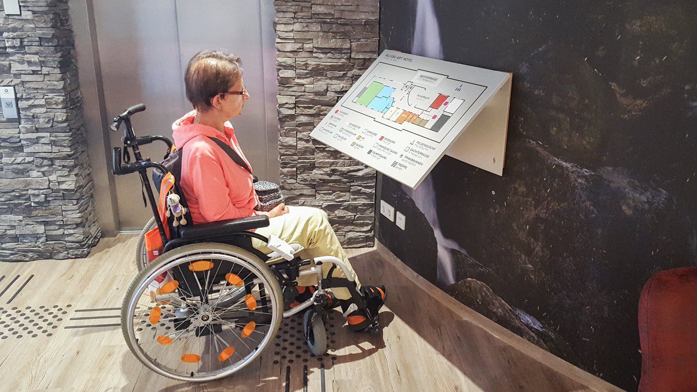 Eine Frau sitzt in einem Rollstuhl vor einem Lageplan. Auf dem Plan sind verschiedene Räume in unterschiedlichen Farben gekennzeichnet. Unterhalb des Plans sind die Farben jeweils beschriftet. Die Beschriftungen sind in Brailleschrift übersetzt. Der Plan kann über ein Bodenleitsystem erreicht werden.