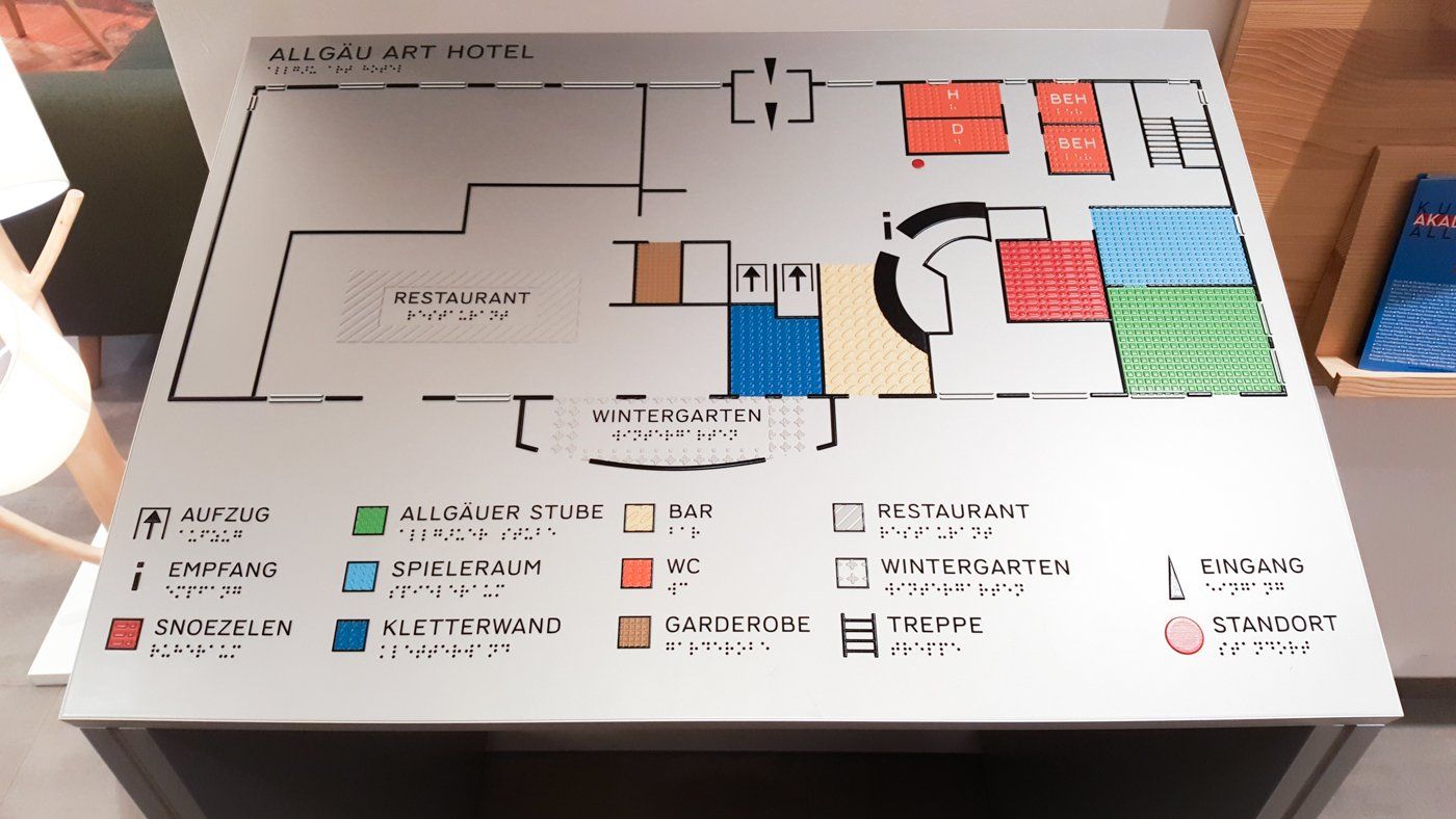 Ein Lageplan des Allgäu Art Hotel, den man mit den Händen erkunden kann. Die Räume sind mit verschiedenen, gut erkennbaren Farben markiert, und es gibt eine Legende, die anzeigt, wo sich die Räume befinden. Diese Informationen sind sowohl im lateinischen Alphabet als auch in Brailleschrift verfügbar, um leicht zu erfahren, wo man ist und wie man zu anderen Räumen gelangt.