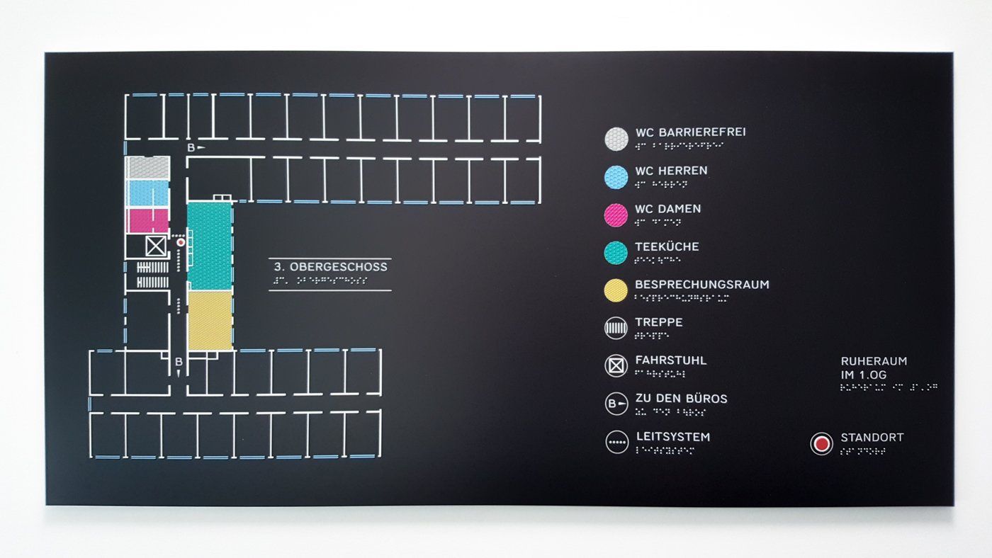 Ein taktiler Lageplan eines Büros. Auf dem Plan sind in die WCs, Teeküche, Büros, Fahrstühle und Treppen eingezeichnet. Die Platte des Plans ist schwarz, die Räume sind in starken, kontrastreichen Farben eingetragen