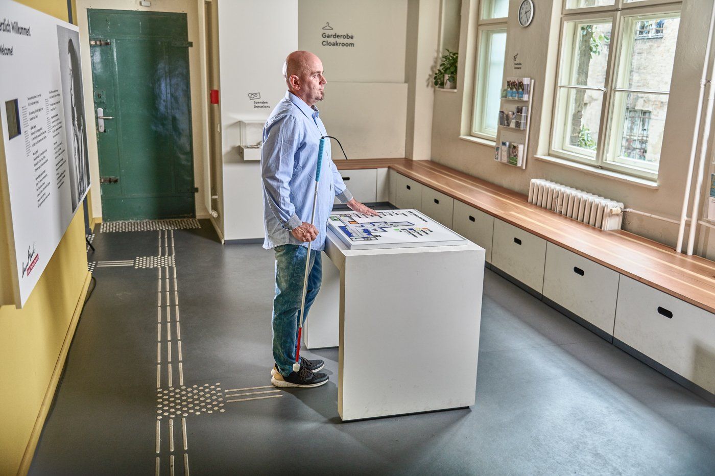 Ein Mann mit Blindenstock steht an einem taktilen Lageplan und ertastet Informationen. Er befindet sich in einer Garderobe. Am Boden befindet sich ein Bodenleitsystem, das von einer Eingangstür zum Plan und weiter in den Raum führt.