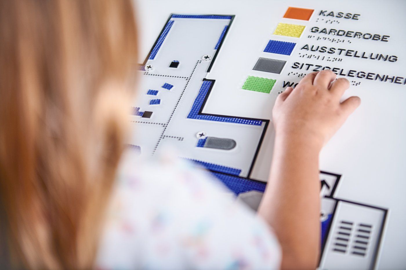 Eine Person steht vor einem taktilen Grundrissplan. Ihre Hand befindet sich über beim Eintrag Sitzgelegenheiten und ertastet das Wort in Brailleschrift. Auf dem Plan sind auch die Kasse, Garderobe und Ausstellung eingezeichnet.